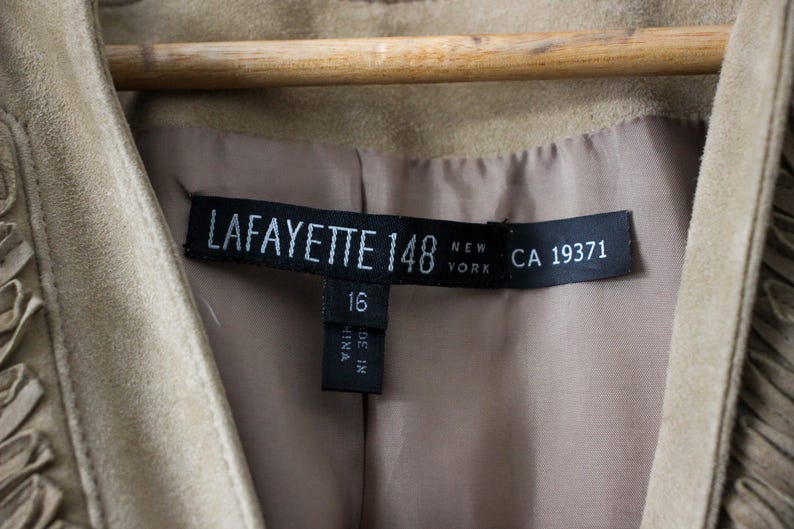May include: Close-up of a tan suede jacket with a label that reads "LAFAYETTE 148 NEW YORK CA 19371". The jacket is hanging on a wooden hanger. The label also indicates size 16.