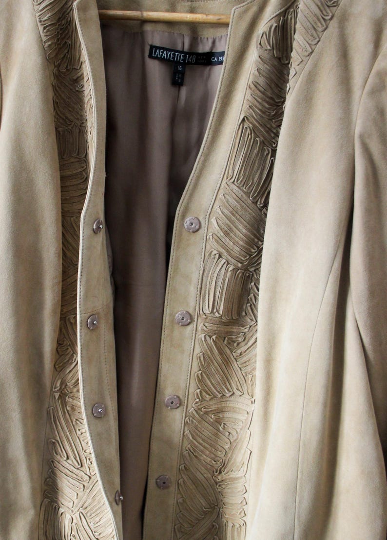 May include: A tan suede jacket with decorative stitching along the front and collar. The jacket has a button-up closure and a soft, neutral color, perfect for layering. The label reads "LAFAYETTE 148".