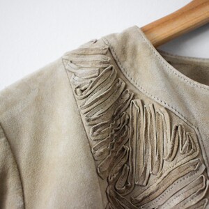 May include: Close-up of a tan suede jacket with decorative fringing along the neckline. The jacket is hanging on a wooden hanger. The neutral color and soft texture suggest a vintage or bohemian style.