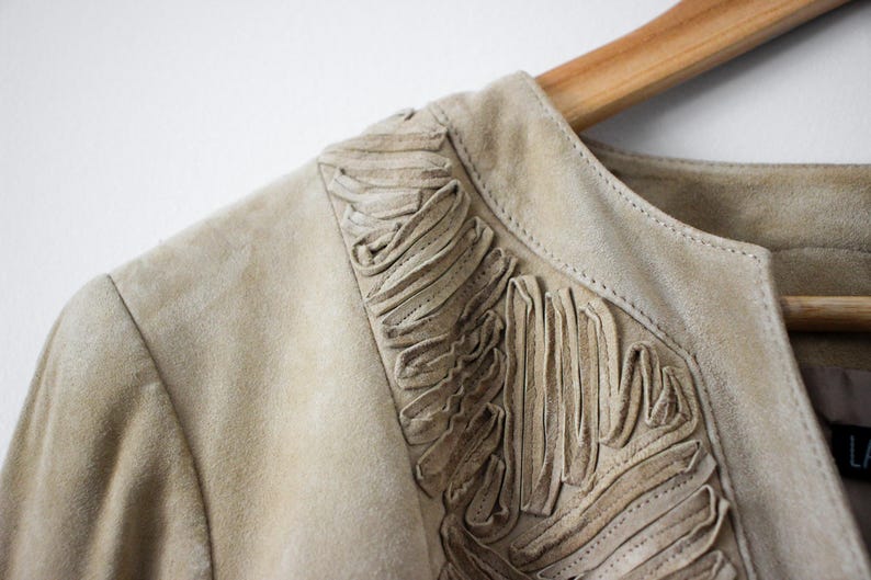 May include: Close-up of a tan suede jacket with decorative fringing along the neckline. The jacket is hanging on a wooden hanger. The neutral color and soft texture suggest a vintage or bohemian style.