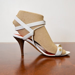 Peut inclure: Une sandale à talons hauts blanche dotée d'une fine bride à la cheville, d'un talon aiguille marron et d'un ornement métallique doré sur la bride avant. La semelle intérieure est rouge et la chaussure est présentée sur un embauchoir beige.
