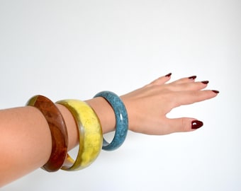 May include: Three colorful resin bangles on a person's wrist. The bangles are yellow, brown, and blue.