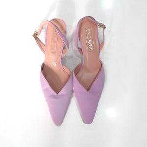 以下が含まれることがあります： 先のとがったつま先と足首ストラップが付いた、ラベンダー色のサテン製ヒールシューズのペア。シューズにはゴールドのバックルが付いており、ストラップには「ESCADA」というブランド名が印刷されています。