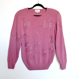 May include: Mauve-coloured jumper with long sleeves and a crew neck. The jumper has a pattern of silver sequin spirals and lines across the front and shoulders. The garment is hanging on a black hanger against a white background.