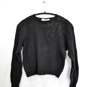May include: A black crew neck sweater with long sleeves, suspended from a hanger. The sweater features a beaded embellishment on the shoulder. The brand name "Aficionados" is visible on the neckline.