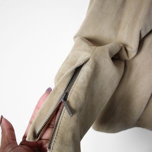 May include: Close-up of a tan suede jacket sleeve with a silver zipper detail. The zipper is partially open, revealing the interior lining. The jacket appears to be made of soft, high-quality material.