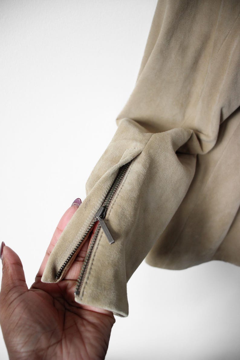 May include: Close-up of a tan suede jacket sleeve with a silver zipper detail. The zipper is partially open, revealing the interior lining. The jacket appears to be made of soft, high-quality material.