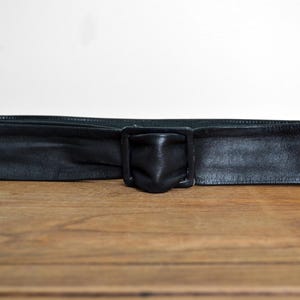 May include: A wide black leather belt with a square buckle. The belt has a pointed end. The buckle is also black and square. The belt is on a wooden surface.
