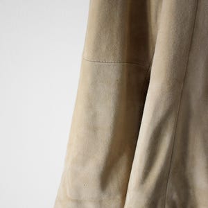 May include: Close-up of a tan suede jacket. The jacket has a soft, textured appearance with visible seams and a curved hem. The color is a light, neutral beige.