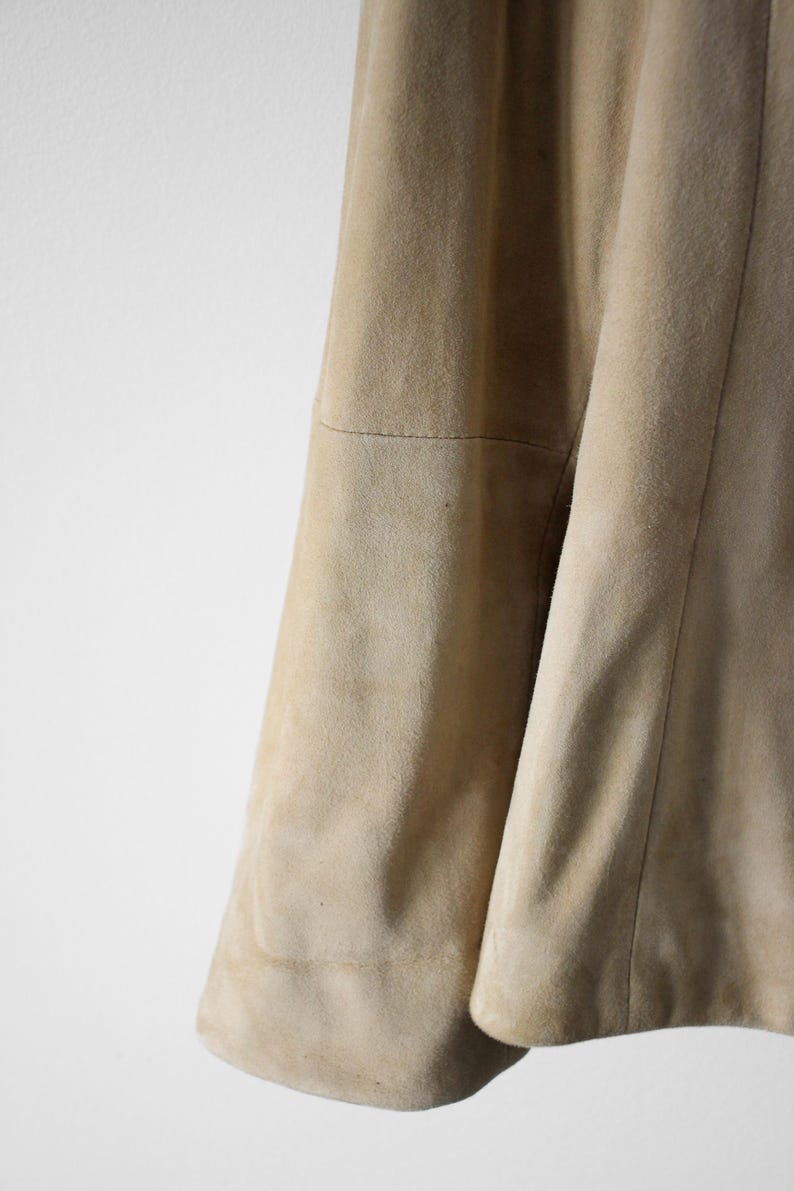 May include: Close-up of a tan suede jacket. The jacket has a soft, textured appearance with visible seams and a curved hem. The color is a light, neutral beige.