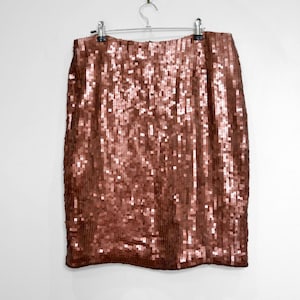 Peut inclure: Jupe à sequins de couleur cuivrée suspendue à un cintre en métal sur fond blanc. La jupe est recouverte de petits sequins rectangulaires qui captent la lumière, créant un effet scintillant. La jupe semble arriver au genou.