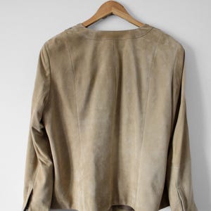 May include: A tan suede jacket hanging on a wooden hanger. The jacket has long sleeves, a collarless neckline, and a zipper detail on the cuffs. The back of the jacket is visible, showing the seams and tailoring.