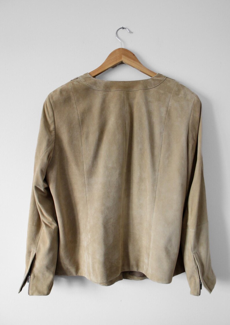 May include: A tan suede jacket hanging on a wooden hanger. The jacket has long sleeves, a collarless neckline, and a zipper detail on the cuffs. The back of the jacket is visible, showing the seams and tailoring.