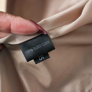 May include: Close-up of a black label with white text that reads "100% SUEDE/SUEDE" and the number "35". The label is attached to a beige fabric. The fabric is held by a hand with red nail polish.