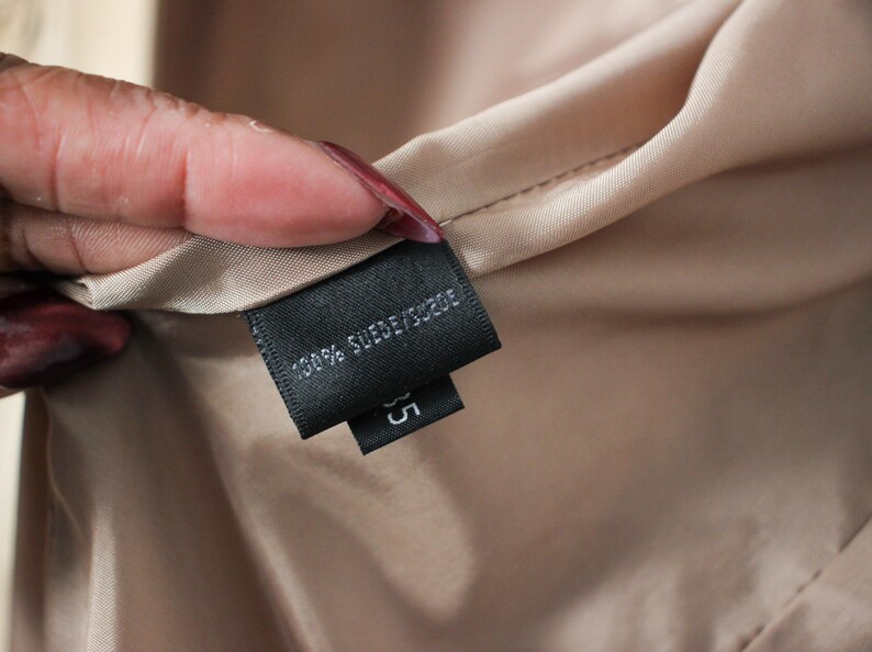 May include: Close-up of a black label with white text that reads "100% SUEDE/SUEDE" and the number "35". The label is attached to a beige fabric. The fabric is held by a hand with red nail polish.