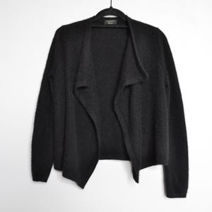 Peut inclure: Un cardigan noir à devant ouvert est suspendu à un cintre noir sur fond blanc. Le cardigan a un col drapé et des manches longues. Le tissu semble être un tricot doux et texturé. Cette pièce polyvalente est idéale pour superposer.