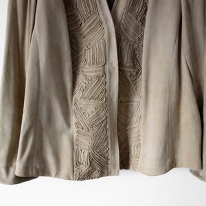May include: A tan suede jacket with a unique textured design down the front. The jacket has long sleeves and a classic open front. The textured detail is a series of raised, geometric patterns. The jacket is a neutral color.