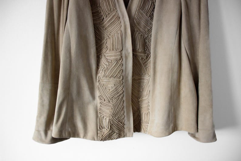 May include: A tan suede jacket with a unique textured design down the front. The jacket has long sleeves and a classic open front. The textured detail is a series of raised, geometric patterns. The jacket is a neutral color.