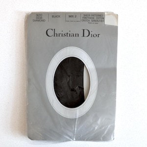 May include: Sealed package of black Christian Dior sheer patterned pantyhose, size 2. The packaging is grey with the brand name and product details printed in black. The pantyhose are visible through an oval window.