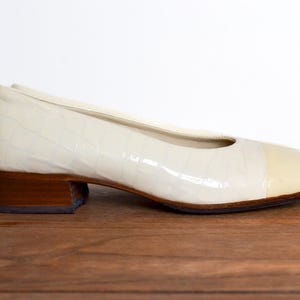 May include: Cream-coloured, low-heeled shoe with a textured, crocodile-like pattern. The toe cap is a lighter shade of cream. The heel is a dark brown, rectangular block. The shoe is set on a wooden surface.