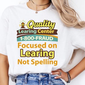 May include: White t-shirt with a humorous graphic. The design includes the words "Quality Learing Center" and "1-800-FRAUD", with the tagline "Focused on Learing Not Spelling". The text is in various colors, including yellow, brown, and teal.