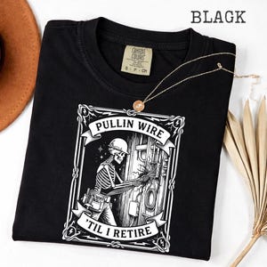May include: Black t-shirt with a graphic of a skeleton electrician working on a panel. The design includes the text "PULLIN WIRE" and "'TIL I RETIRE" in a decorative frame. The shirt has a crew neck and short sleeves.