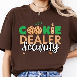 May include: A brown t-shirt with the words "COOKIE DEALER SECURITY" in green, tan, and white. The word "COOKIE" features two cookie graphics. The shirt is a solid brown color.