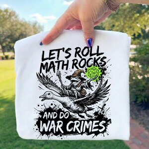 May include: White sweatshirt with a black and white graphic of a frog wizard riding a goose, holding a glowing green die. Text reads "LET'S ROLL MATH ROCKS AND DO WAR CRIMES."