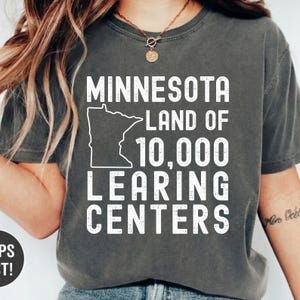 May include: Dark gray t-shirt with the text "MINNESOTA LAND OF 10,000 LEARNING CENTERS" in white. The design includes a white outline of the state of Minnesota. The shirt is a casual wear item.
