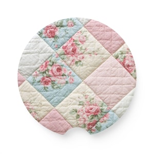 May include: A circular car coaster featuring a quilted diamond pattern with alternating squares of solid light pink, cream, and a floral print displaying pink roses on a light blue background. The coaster has a small semi-circular notch at the bottom.
