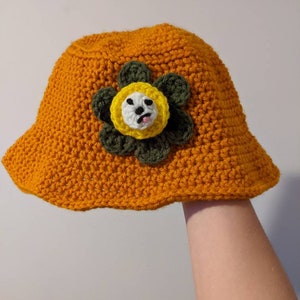 May include: A crocheted bucket hat in orange with a green and yellow flower appliqué. The flower has a white face with a black mouth and eyes.