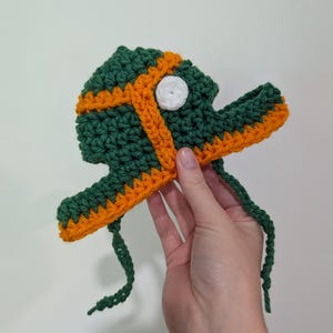May include: A handmade crochet fish hat in green, orange, and white. The hat features a green body with orange trim and a white eye. Two green ties extend from the hat.