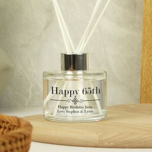 Puede incluir: Una botella de difusor de vidrio transparente con una tapa plateada y texto negro que dice "Happy 65th" y "Happy Birthday June Love Stephen & Lynn".