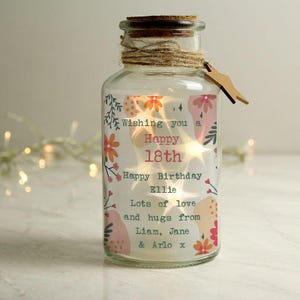 May include: A clear glass jar with a cork stopper, twine, and a wooden tag. The jar is decorated with floral designs and text that reads "Wishing you a Happy 18th Happy Birthday Ellie Lots of love and hugs from Liam, Jane & Arlo x."