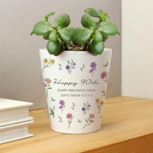 May include: A white ceramic planter with a green succulent plant. The planter is decorated with colorful floral illustrations and the text "Happy 90th Happy Birthday Gran Love From Lucy X". The planter sits on a wooden surface.
