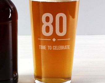 May include: A clear glass pint glass with the number 80 etched on the front. The text "Time to Celebrate" is below the number. The glass is filled with a golden-colored beverage.