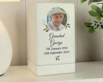 May include: White wooden box with a photo of a man in a cap. The box has a gold trim and a floral design. The text on the box reads "Grandad George 7th January 1934 12th September 2025".