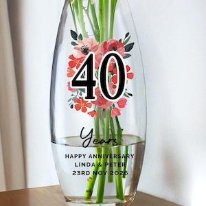Peut inclure: Un vase en verre transparent avec un motif floral et le chiffre "40" en noir. Le vase contient de l'eau et des tiges vertes. Le texte sur le vase indique "Years" et "Happy Anniversary Linda & Peter 23rd Nov 2026".