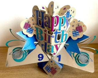 May include: A pop-up card with a blue, pink, and purple "Happy Birthday" message. The card is decorated with silver glitter and blue stars. The card is open to reveal the numbers "9" and "1" in silver glitter.