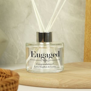 Puede incluir: Una botella difusora de vidrio transparente con una tapa plateada y cañas blancas. La botella tiene una etiqueta negra que dice "Engaged" con un diseño decorativo y el texto "¡Felicidades! Con cariño, Stephen y Lynn".