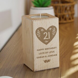 May include: A wooden candle holder with a heart-shaped design featuring the number 21 and botanical accents. The candle holder has the text "HAPPY BIRTHDAY LOTS OF LOVE SARAH & JORDAN" engraved on the front. A lit candle sits inside.
