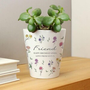 May include: A white ceramic plant pot with a green succulent. The pot is adorned with colourful floral designs and the word "Friend". Text reads "Happy Birthday Linda Love From Lucy X".