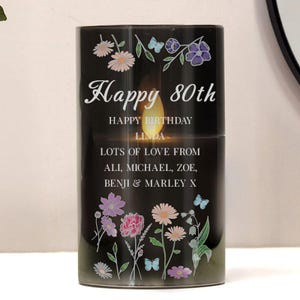 Op de afbeelding: Een zwarte kaars met de tekst "Happy 80th Happy Birthday Linda Lots of Love From Ali, Michael, Zoe, Benji & Marley X". De kaars is versierd met bloemen- en vlinderontwerpen in roze, paars en wit.