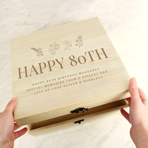 May include: A light-coloured wooden box with the words "HAPPY 80TH" engraved on the lid, along with floral designs. The box is open, revealing a drawer. Text below reads "HAPPY 80TH BIRTHDAY MARGARET SPECIAL MEMORIES FROM A SPECIAL DAY LOTS OF LOVE OLIVER & SCARLETT."