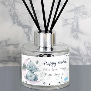 Op de afbeelding: Transparante glazen diffuserfles met zwarte stokjes. Het etiket toont een grijze teddybeer op een maan, met de tekst "Happy 65th With love Mum From Amy". Sterren en bloemmotieven omringen de tekst.