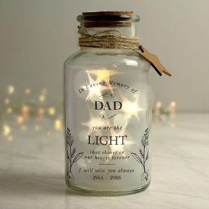 May include: A clear glass jar with a cork stopper, wrapped in twine with a small wooden star tag. Inside, warm fairy lights glow. The front features black text: In Loving Memory of DAD you are the LIGHT that shines in our hearts forever I will miss you always 1955 - 2026, with floral illustrations.