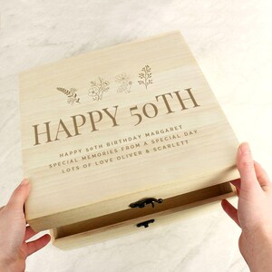 May include: A light-colored wooden box with the words "HAPPY 50TH" engraved on the lid, along with floral designs. The box is open, revealing a drawer. Text below reads "HAPPY 50TH BIRTHDAY MARGARET SPECIAL MEMORIES FROM A SPECIAL DAY LOTS OF LOVE OLIVER & SCARLETT."
