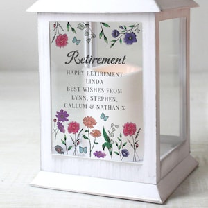 May include: White lantern with a glass panel featuring a floral design and the text "Retirement, Happy Retirement Linda, Best Wishes From Lynn, Stephen, Callum & Nathan X".