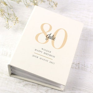 Op de afbeelding: Een wit fotoalbum met een gouden inscriptie die luidt: "80 Julie Walker Gefeliciteerd met je verjaardag 26 augustus 2023".