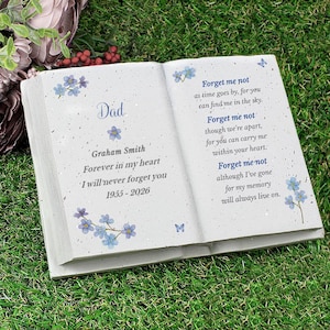 Op de afbeelding: Een witte, open gedenksteen in de vorm van een boek met blauwe bloemaccenten en de woorden "Papa" en "Vergeet me niet" gegraveerd. De tekst bevat een naam, data en een oprechte boodschap, geschikt voor herdenking.